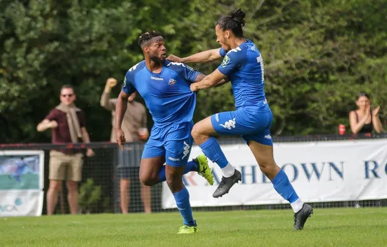 Two footballers in blue celebrating a goal in front of a handful of supporters at a non-league ground