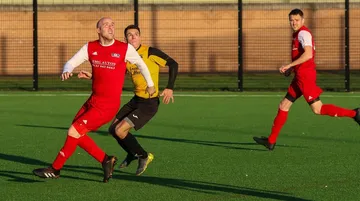 Two adult footballers, one in red and one in yellow, tracking a ball on an artificial pitch in low winter sun