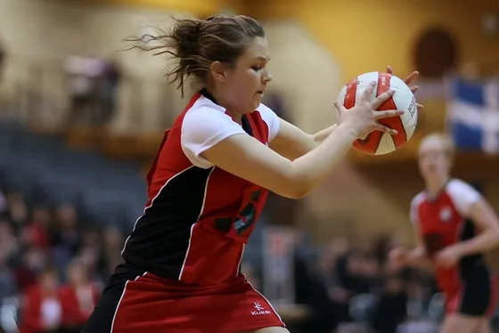 A netball player in red catching the ball at full stretch, a teammate behind her