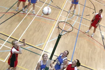 Netball players seen from above in a sports hall, looking up at the ball as it drops towards the ring
