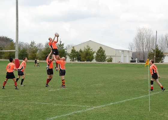 A rugby lineout on a club pitch: a player in orange lifted high to take the ball, teammates waiting